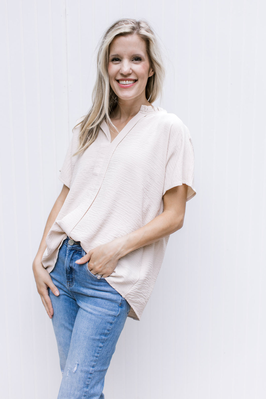 Model wearing jeans with an oatmeal short sleeve top with a v-neck and a pleat down the center.