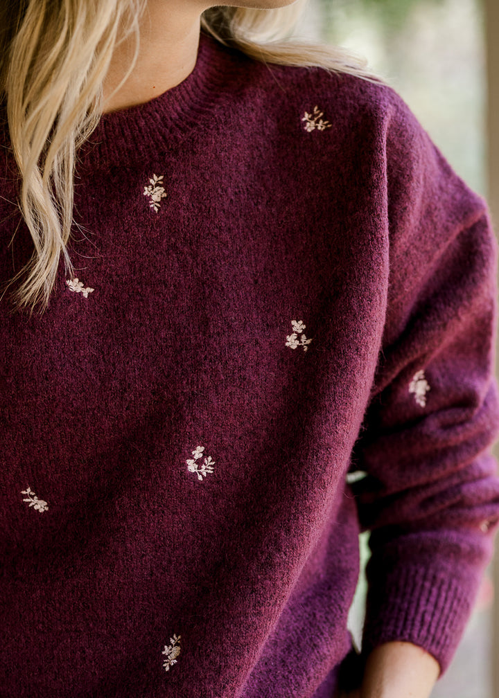 Model wearing a long sleeve plum sweater with cream embroidered flowers and a round neck. 