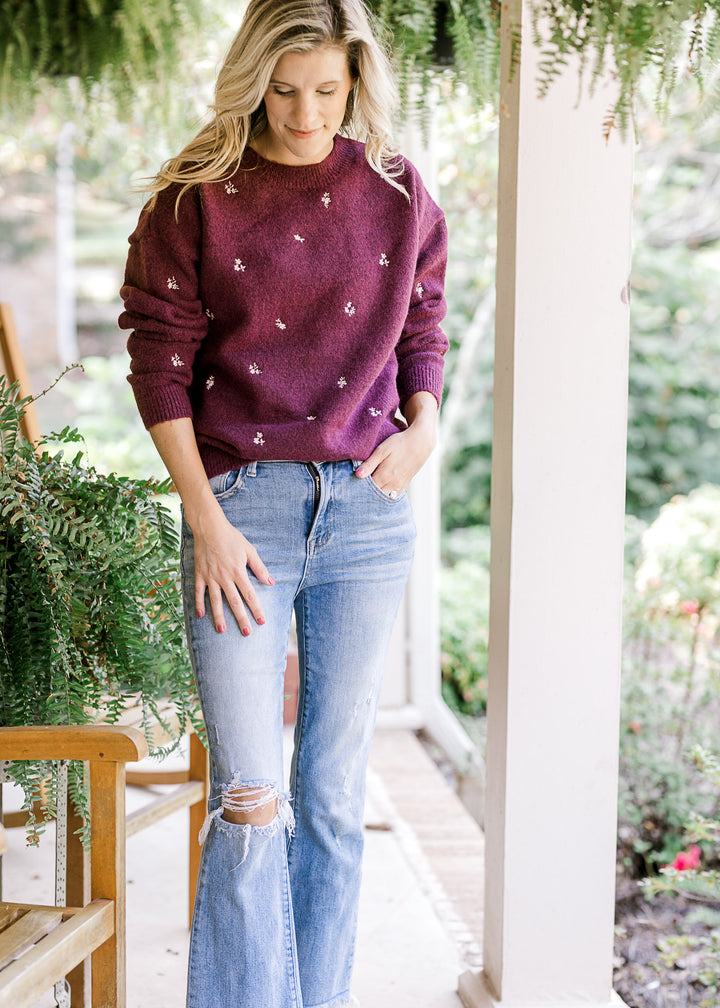 Model wearing jeans and a long sleeve plum sweater with cream flowers and a round neck. 