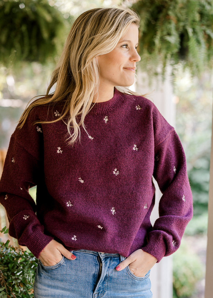 Model wearing a plum sweater with cream embroidered flowers, long sleeves and a round neck. 