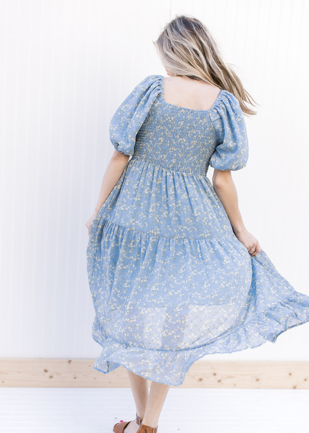 Back view of Model wearing a baby blue dress with white flowers, square neck and short sleeves.