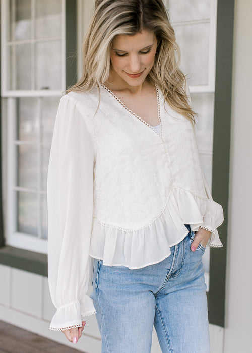 Model wearing a cream top with embroidered bodice, scalloped detail and sheer long sleeves.