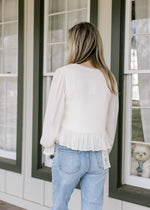 Back view of Model wearing a cream top with scalloped detail, ruffle hem and sheer long sleeves.