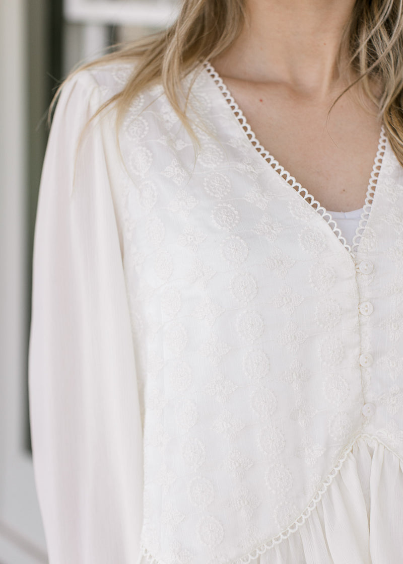 Close up of scalloped hem, embroidered bodice and button closure on a cream long sleeve top.