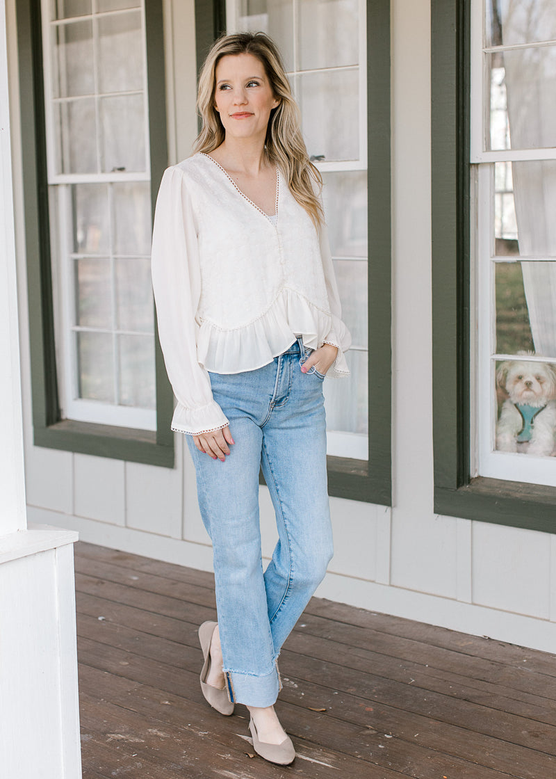 Model wearing jeans and a cream top with embroidered bodice, scalloped detail and long sleeves.