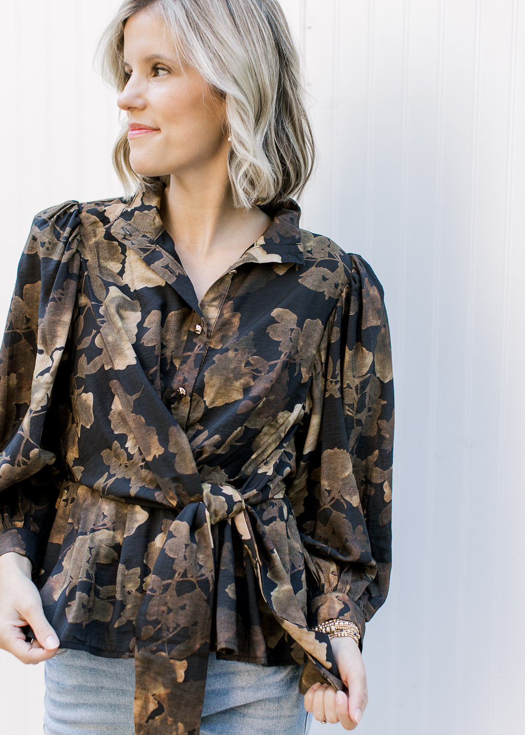 Model wearing a black long sleeve peplum top with brown floral, gold buttons and side tie at waist.