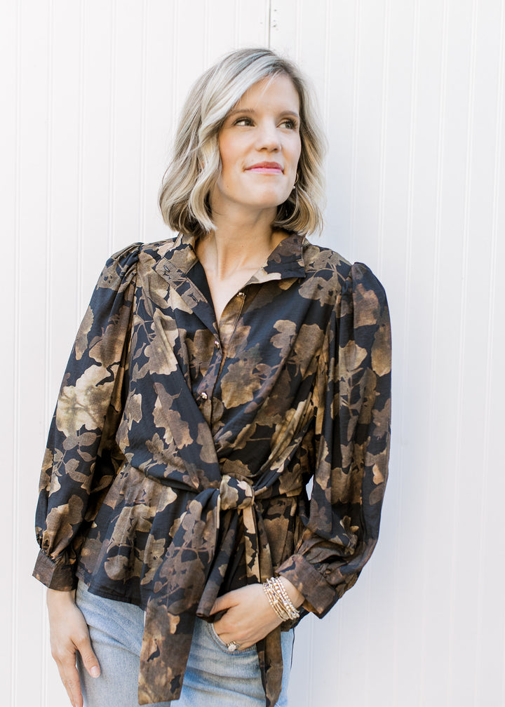 Model wearing a black peplum top with brown floral, gold buttons, side tie and long sleeves. 