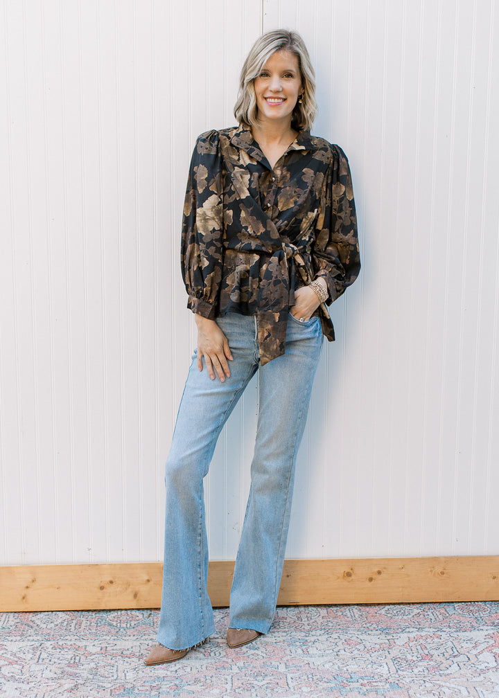 Model wearing jeans, heels and a black top with brown floral print, cold buttons and side tie. 