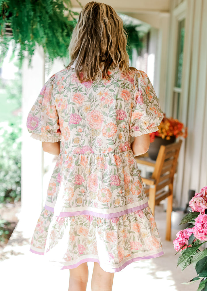 Back view of model wearing a cream dress with floral, short puff sleeves and a tiered skirt. 