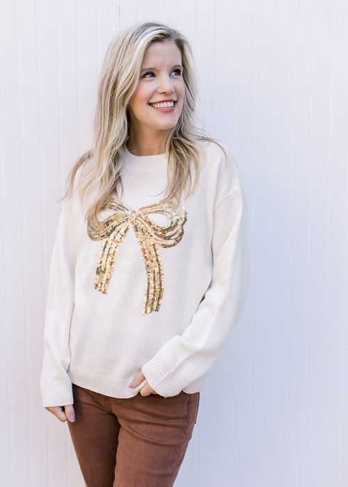 Model wearing a cream sweater with a gold sequin bow across the front and long sleeves.