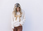 Model wearing a cream sweater with a gold sequin bow, round neck and long sleeves.