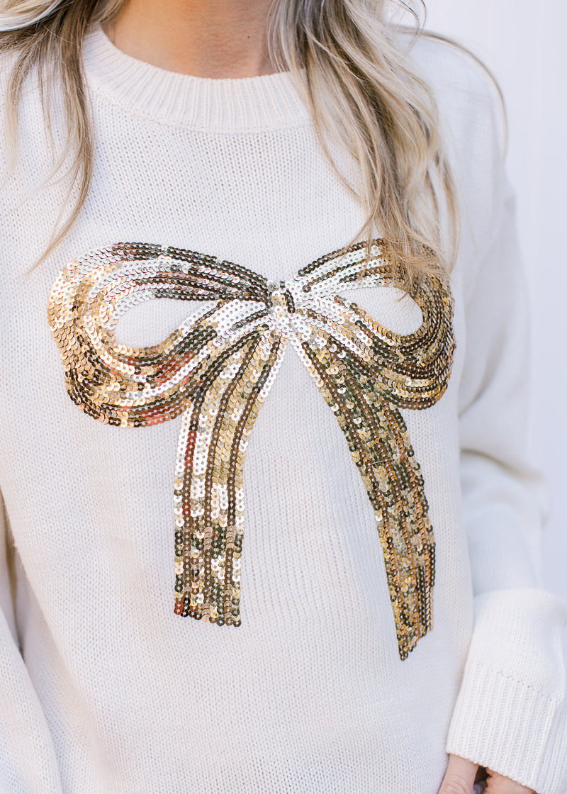 Close up of gold sequin bow on the front of a cream top with long sleeves and a round neck.