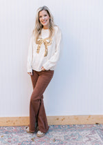 Model wearing espresso pants and a cream sweater with a gold sequin bow and long sleeves.