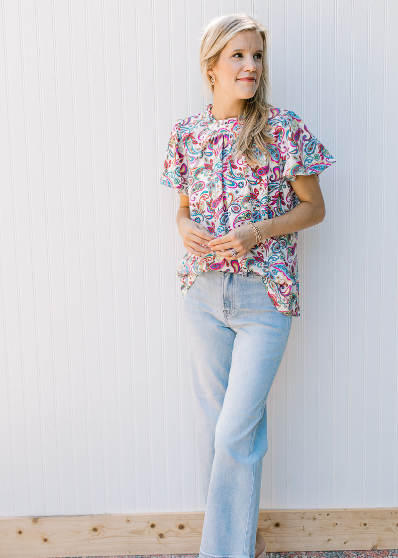 Model wearing a cream top with bright paisley and flutter short sleeves. 