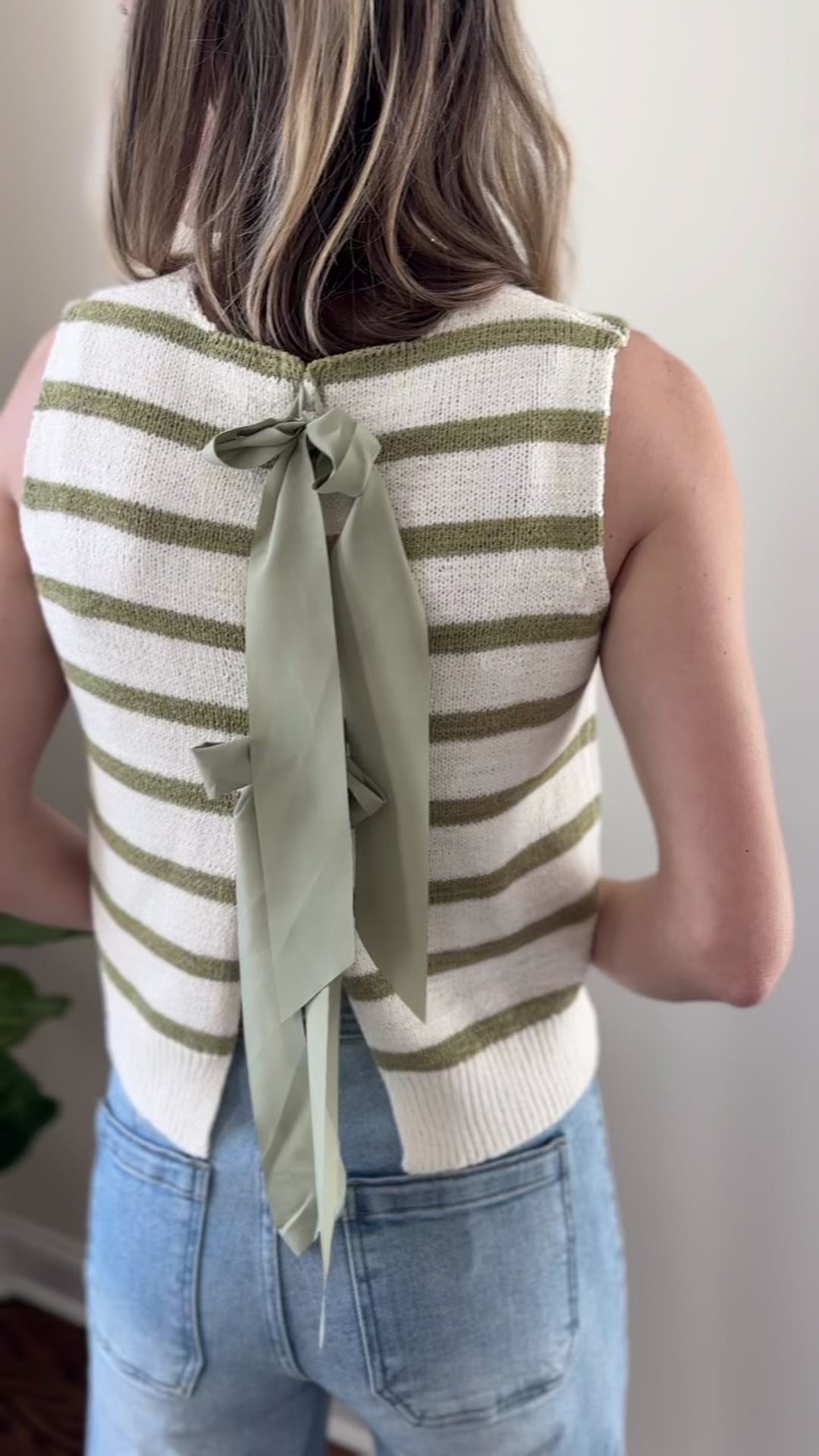 Video of Model wearing a sleeveless cream top with sage stripes and open back with tie closure.