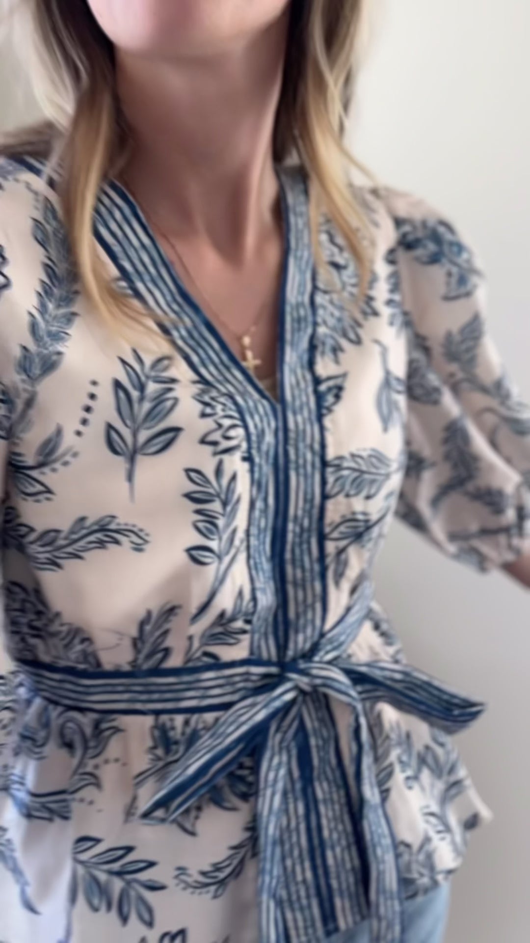 Video of Model wearing a cream v-neck top with blue print and blue stripe detail at placket and waist.