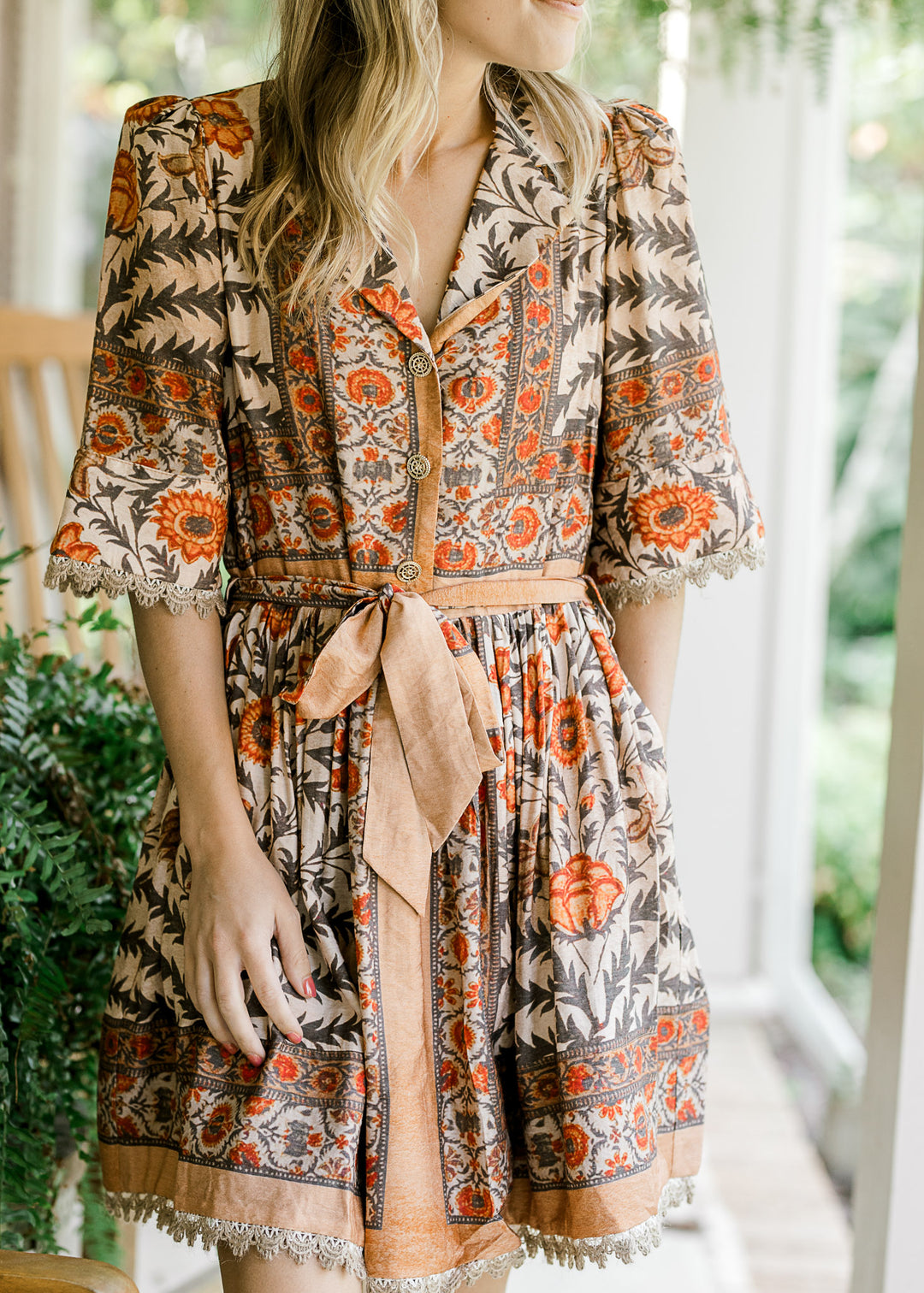Model wearing a tan above the knee dress  with a floral design v-neck, short sleeves and tie waist. 