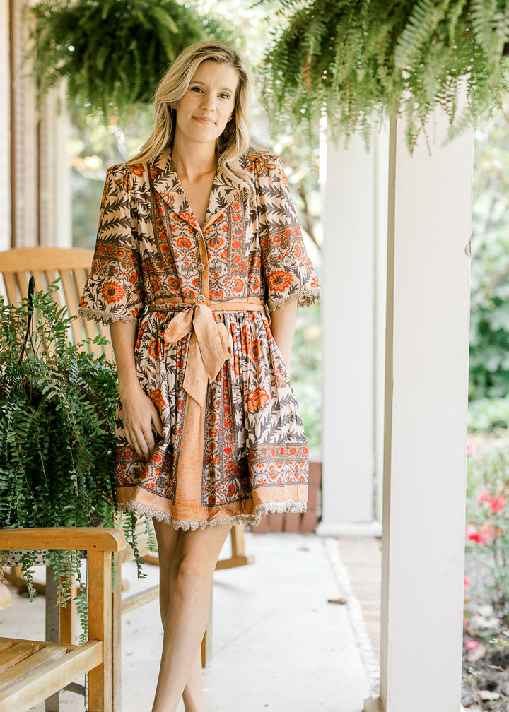 Model wearing a tan above the knee dress  with a floral design, short sleeves and tie waist. 