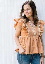 Model wearing a dusty orange top with ruffle cap sleeve and scallop white detail. 