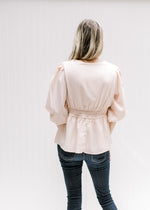 Back view of Model wearing a blush colored top with a cinched waist, v-neck and 3/4 bubble sleeves. 