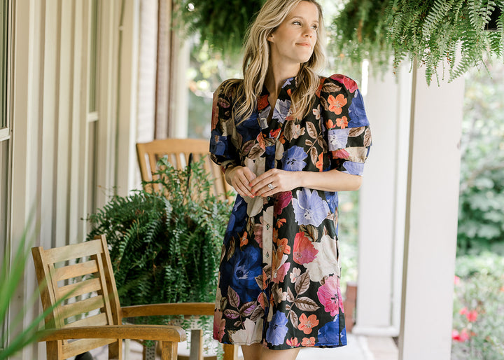 Model wearing an above the knee black dress with floral print, button front and short sleeves. 