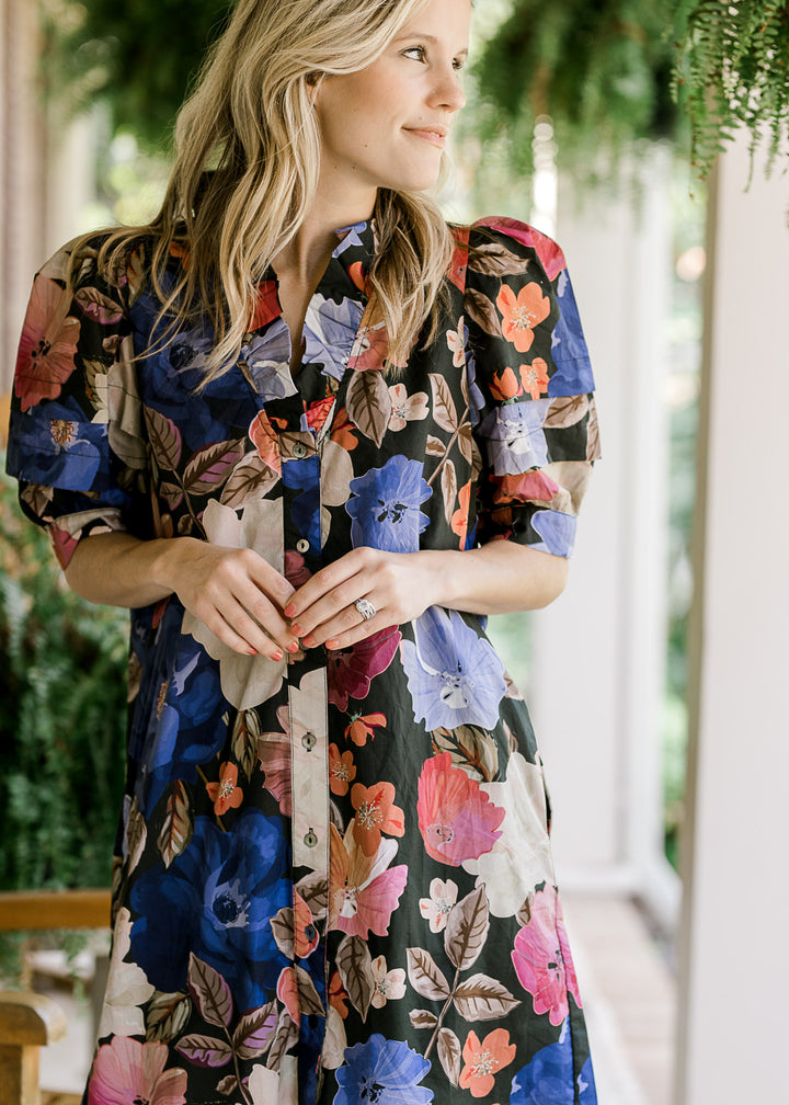 Model wearing a black dress with floral print, v-neck with ruffle, button front and short sleeves. 
