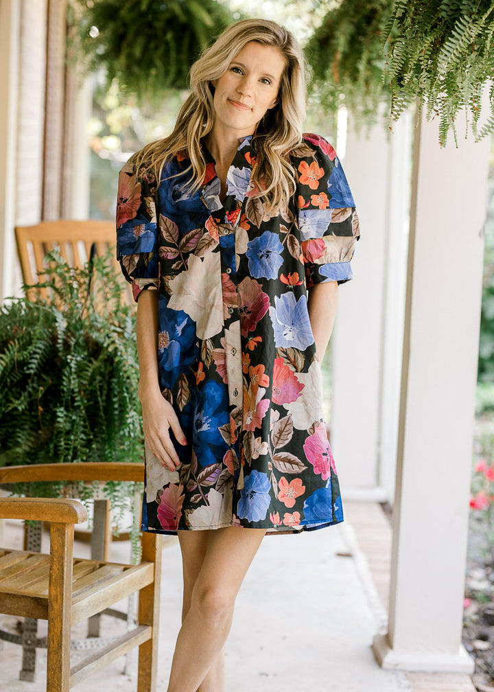 Model wearing a black dress with floral print, pockets, button front and short sleeves. 