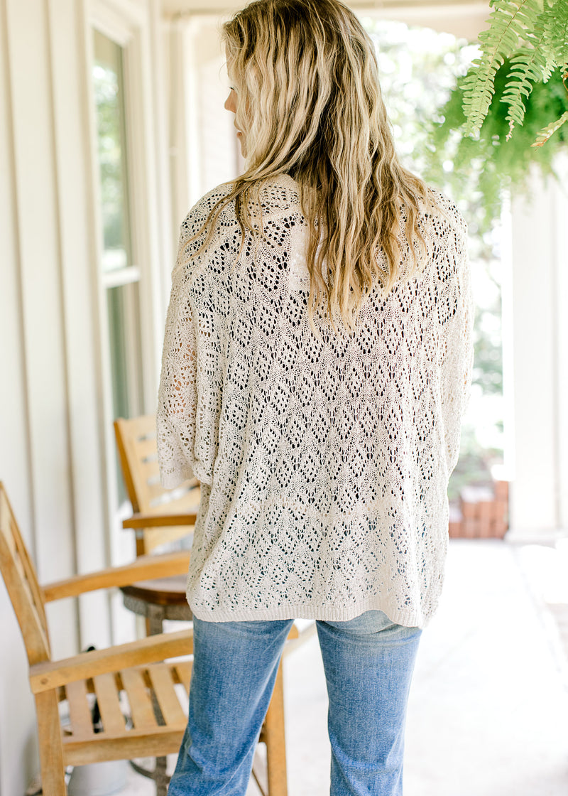 Back view of Model wearing a neutral open weave cardigan with scalloped hem and 3/4 sleeves. 