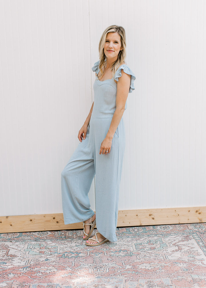 Model wearing sandals and a light blue jumpsuit with ruffle straps, wide legs and a sweetheart neck.