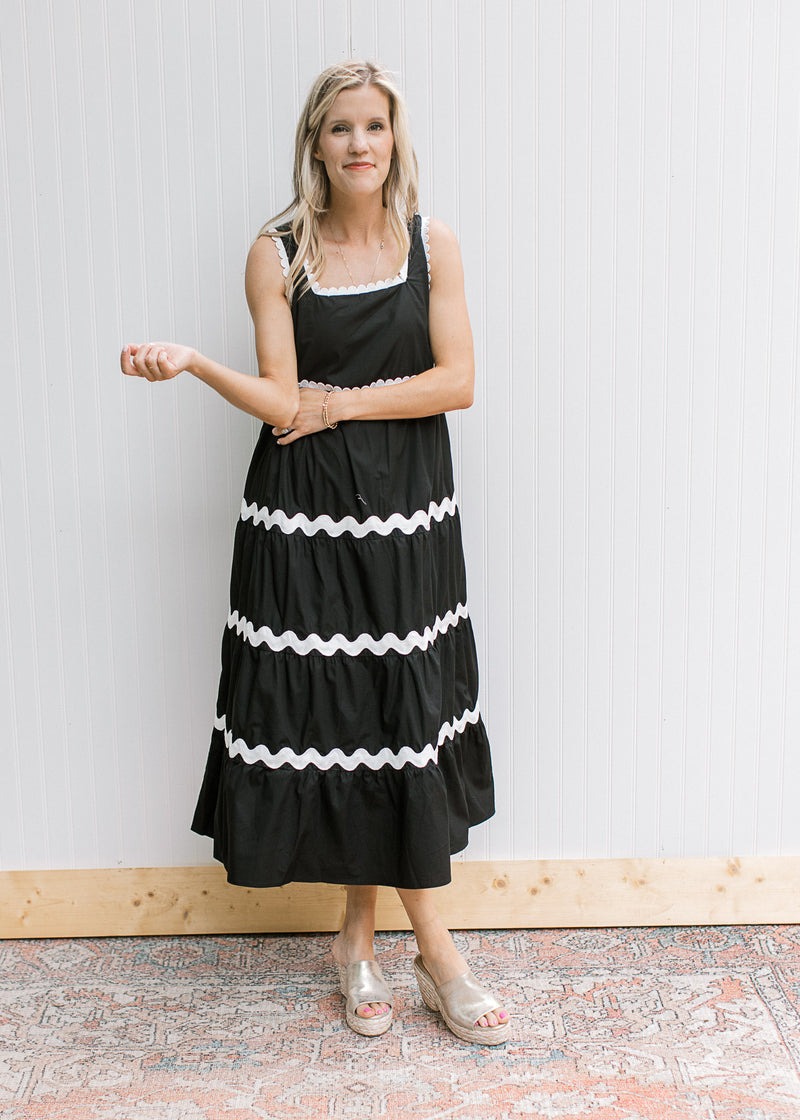 Model wearing a sleeveless black dress tiered with white zig-zag stripe detail and a square neck.