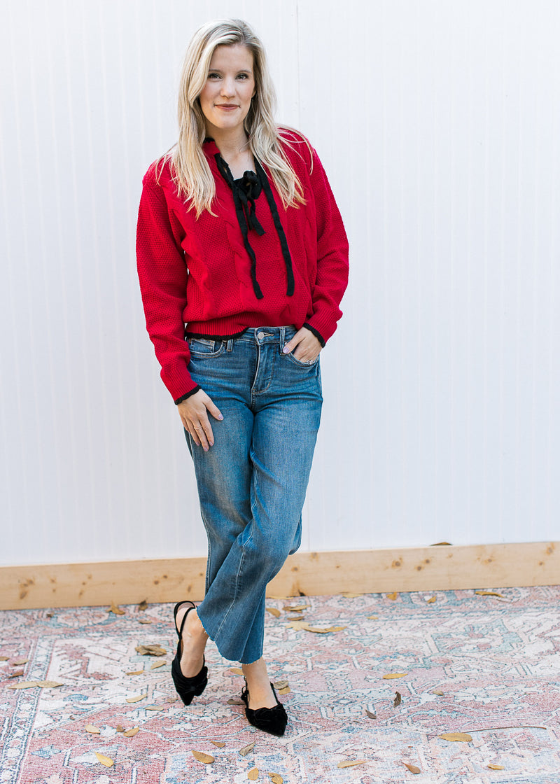 Model wearing jeans, flats and a red knit sweater with black tie bow at v-neck and long sleeves. 