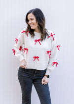 Model wearing a cream sweater adorned with red 3D bows, long sleeves and a round neck. 