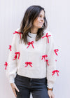 Model wearing a cream sweater adorned with red 3D bows and ribbed detail at neck, cuff and hem.