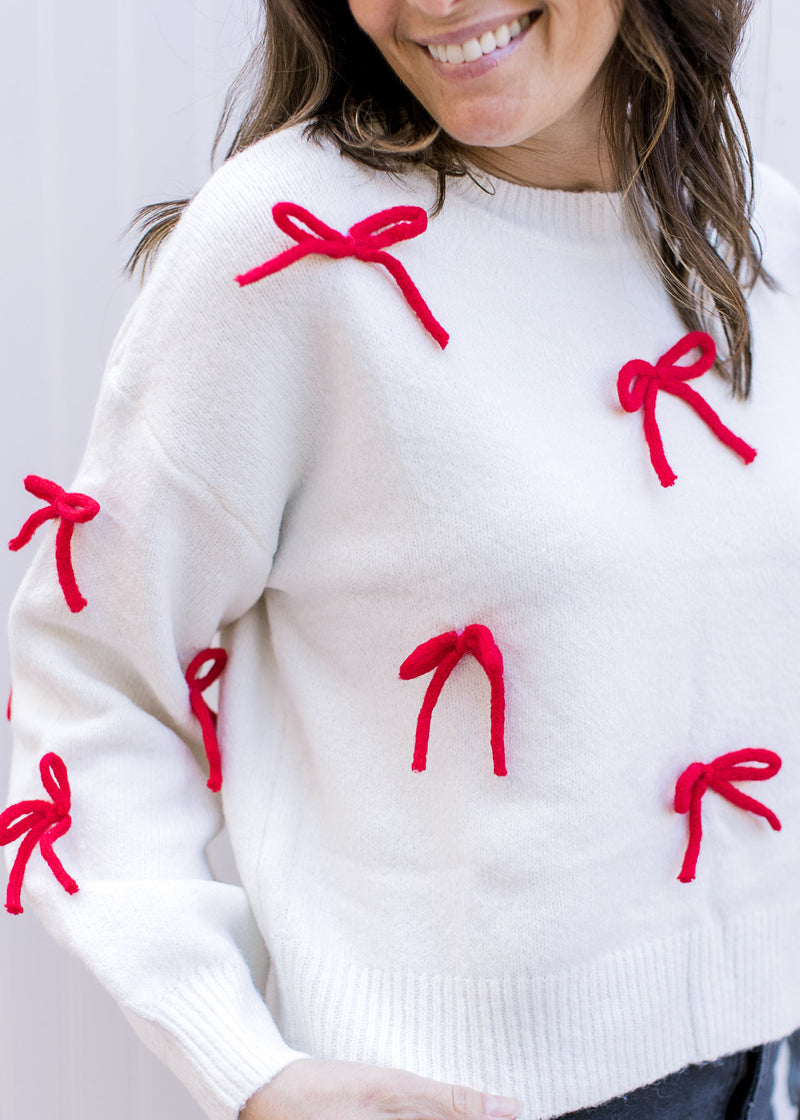 Close up of red 3D bows on a cream sweater with long sleeves and a round neck.