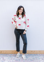 Model wearing black jeans and a cream sweater with red 3D bows, long sleeves and a round neck. 