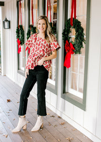 Model wearing black jeans and booties with a cream top with red poppy’s and a high square neck.