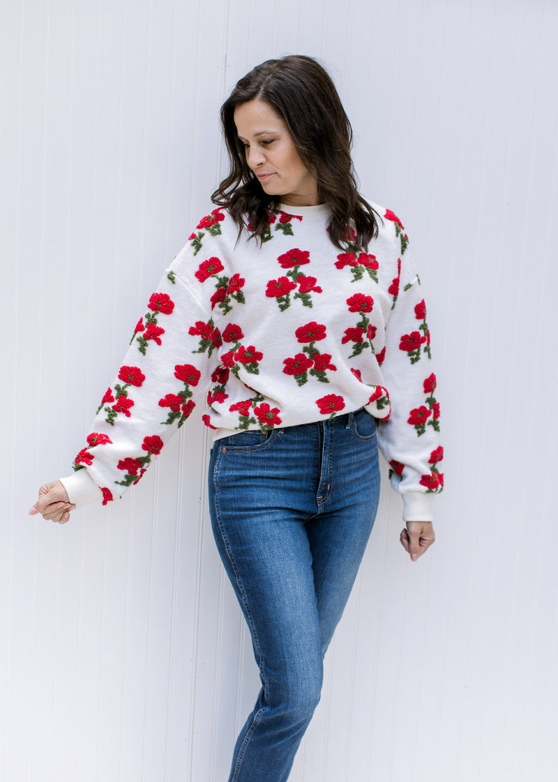 Model wearing jeans and an ivory sweater with textured red poppies, long sleeves and round neckline.