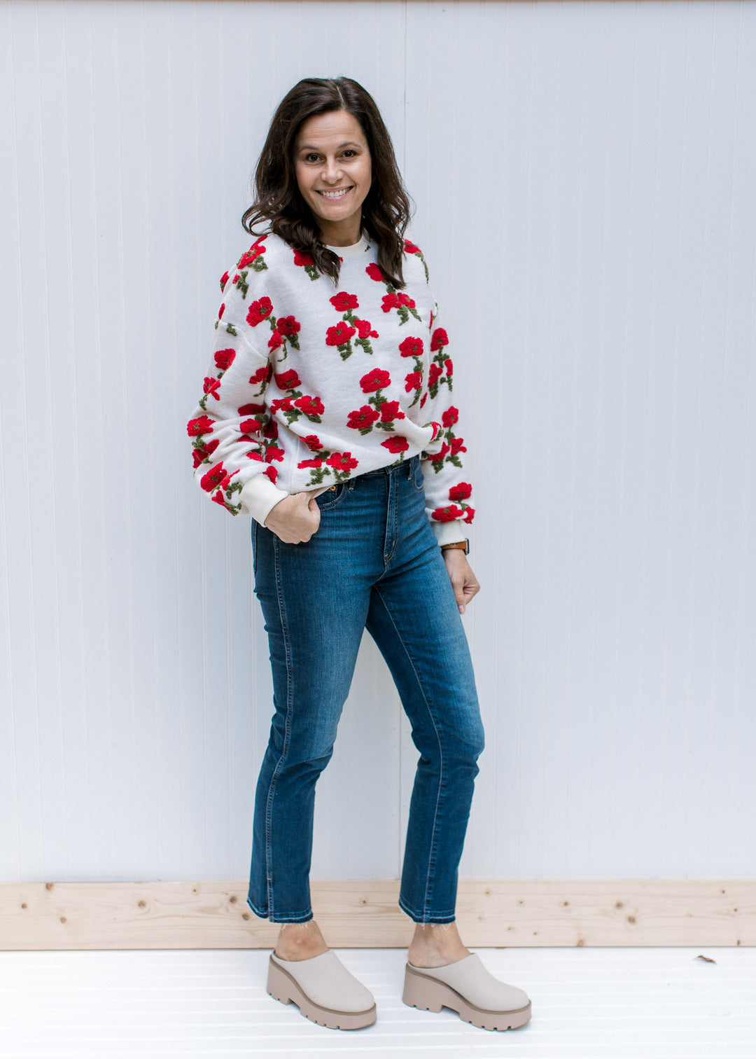 Model wearing jeans, mules and an ivory sweater with red poppies, long sleeves and round neck.