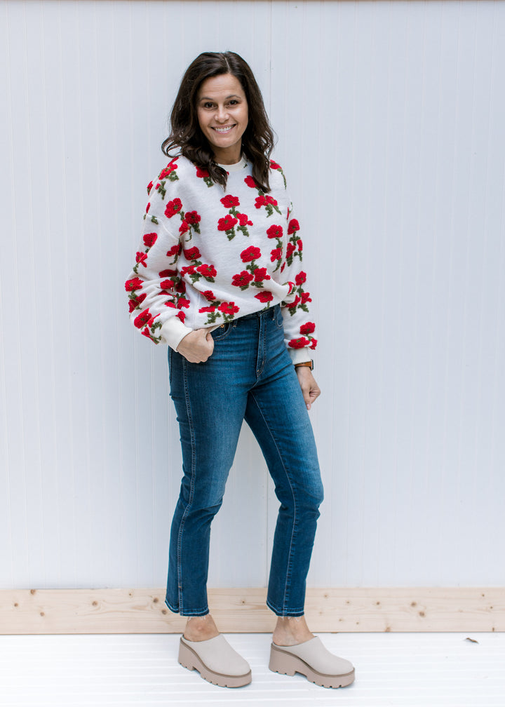 Model wearing jeans, mules and an ivory sweater with red poppies, long sleeves and round neck.