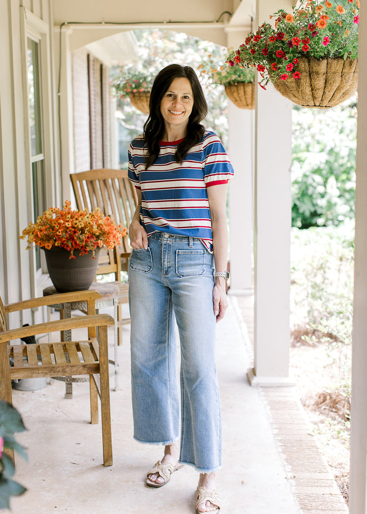 Model in jeans, sandals and a red, cream, and blue striped tee with short sleeves and a round neck. 