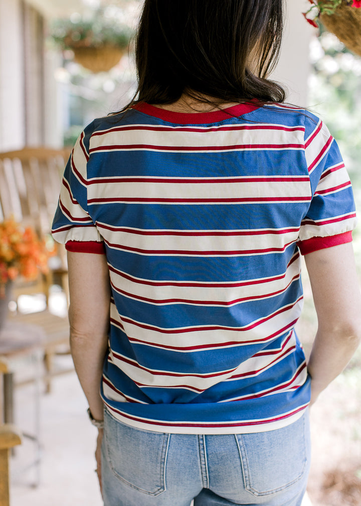 Back view of Model wearing a red, cream, and blue striped tee with short sleeves and a round neck.