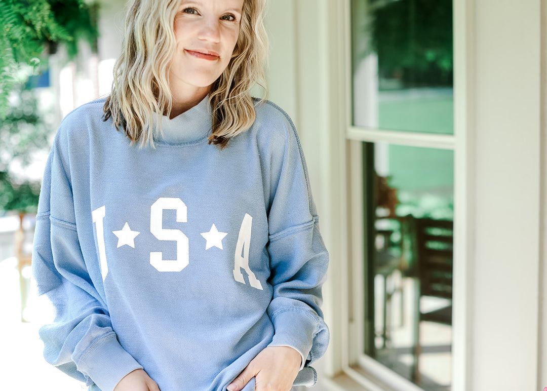 Model wearing a pale blue lightweight sweatshirt with white "USA" graphic and mock neck.