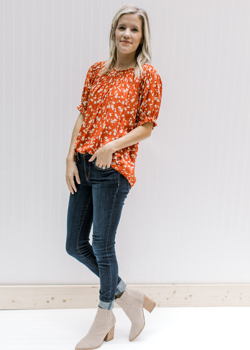 Model wearing jeans, booties and a rust top with cream floral, detail at neck and short sleeves. 