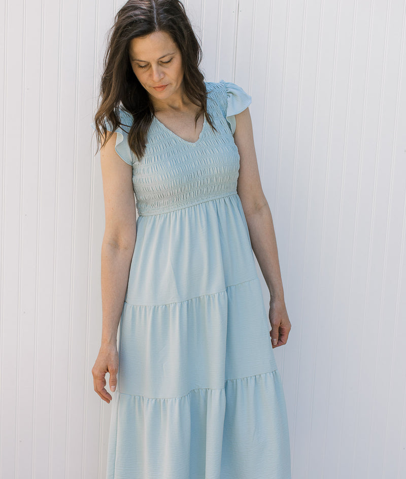 Model wearing a light blue tiered midi with a smocked bodice, v-neck and flutter capped sleeves. 
