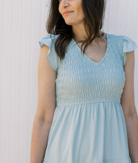 Close up of smocked bodice, v-neck and ruffle capped sleeves on a tiered light blue midi. 