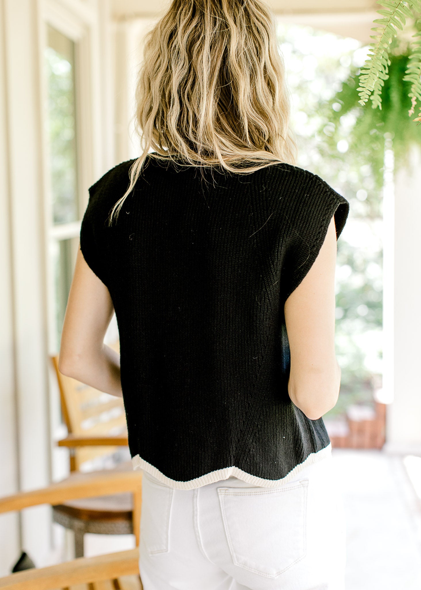 Back view of Model wearing a black ribbed sweater with capped sleeves and cream scalloped hem.