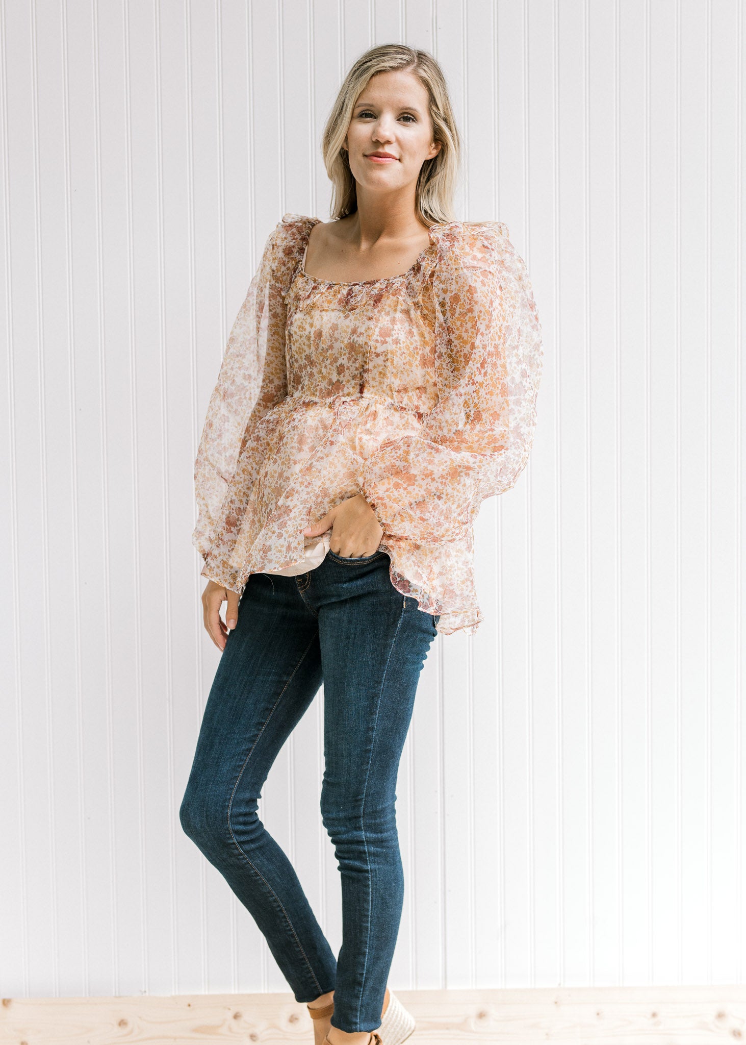 Model wearing jeans, wedges and a beige top with a rust floral pattern and sheer long sleeves. 