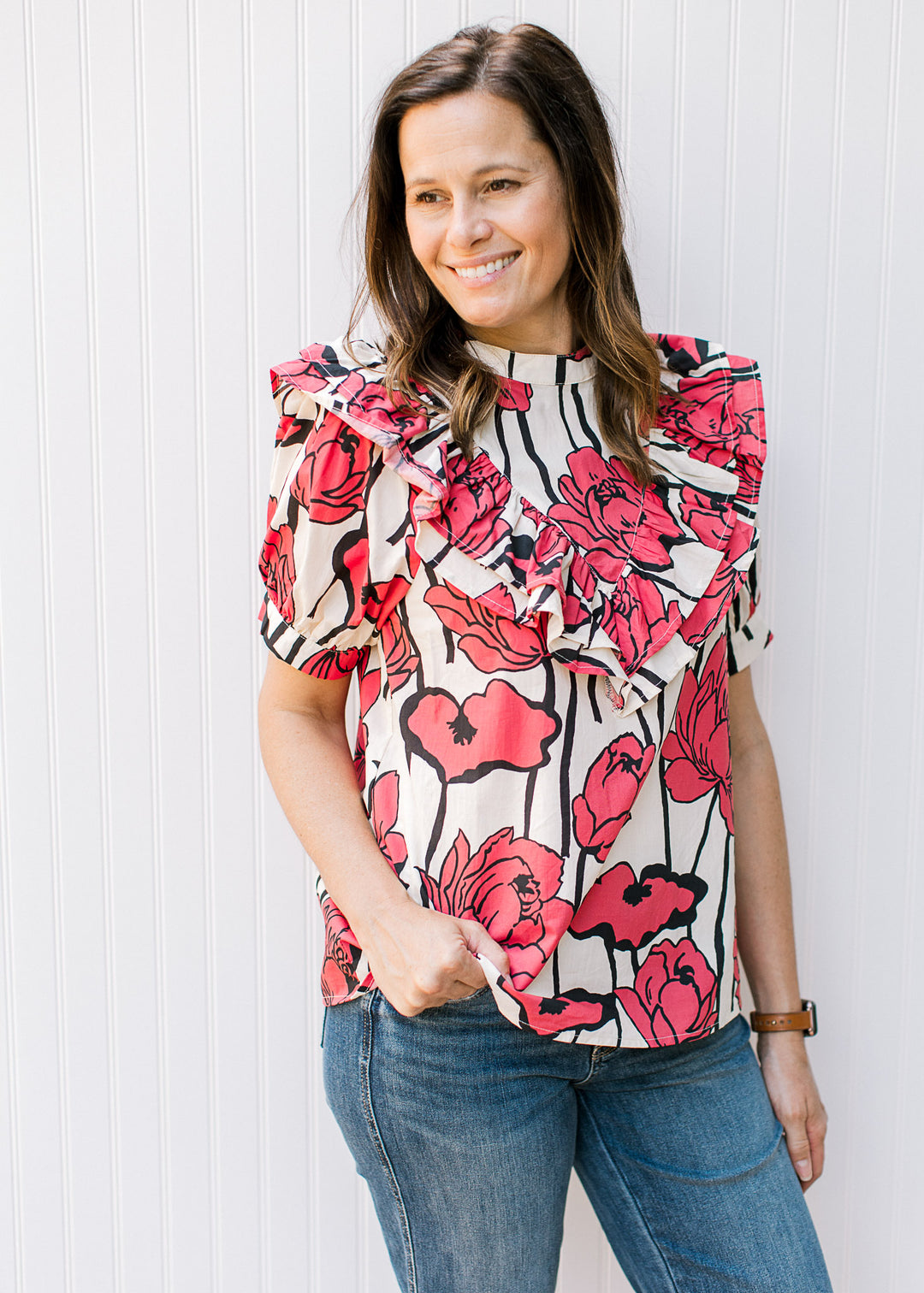 Model wearing a cream top with rose colored flowers, a bib ruffle detail and short sleeves. 
