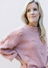 Model wearing a rose colored sweater with a mock neck, 3D flowers and long sleeves. 