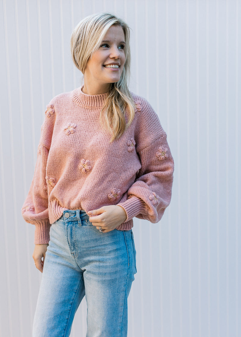 Model wearing jeans with a rose colored sweater with a mock neck, 3D flowers and long sleeves. 